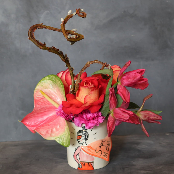 Dark background with arrangement that is a girl with a mask and bun with a cape that says "super MOM". Flowers in the vase are pink tulips, coral roses, onepink  anthurium and two pussy willow branches