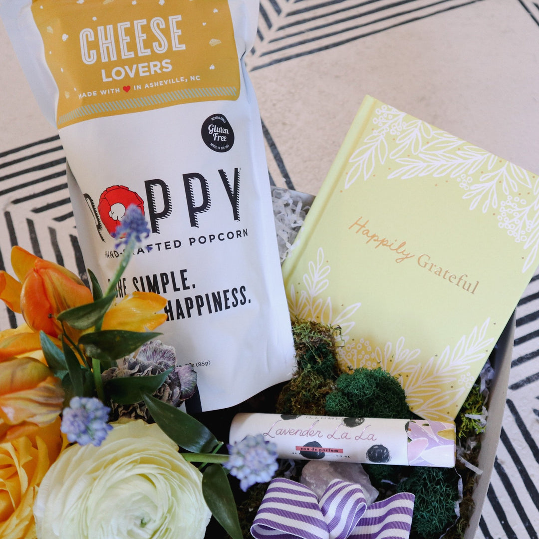 Thank You Gift Box | Close up on the gift box with Poppy Popcorn "Cheese Lovers" Popcorn, a small spring arrangement with yellow, blue, orange, and white, a perfume, and a yellow book titled "Happily Grateful". The box is decorated with white shredded paper and moss.