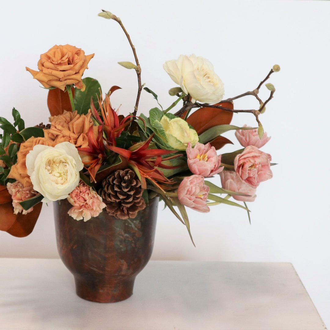 Arrangement in a brown metal vessel, filled with pink tulips, kale, carnations, toffee roses, amaryllis, magnolia branches, pinecones, greenery and magnolia. On a light background.