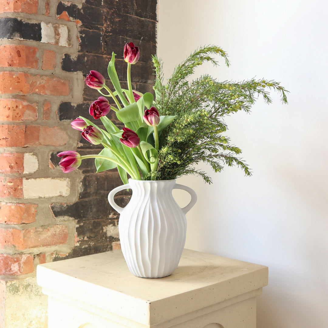 Hand In Hand | A vased arrangement with pink tulips. This arrangement is in a white vase with ribbed sides and a handle on each side. Phot taken against a brick background.