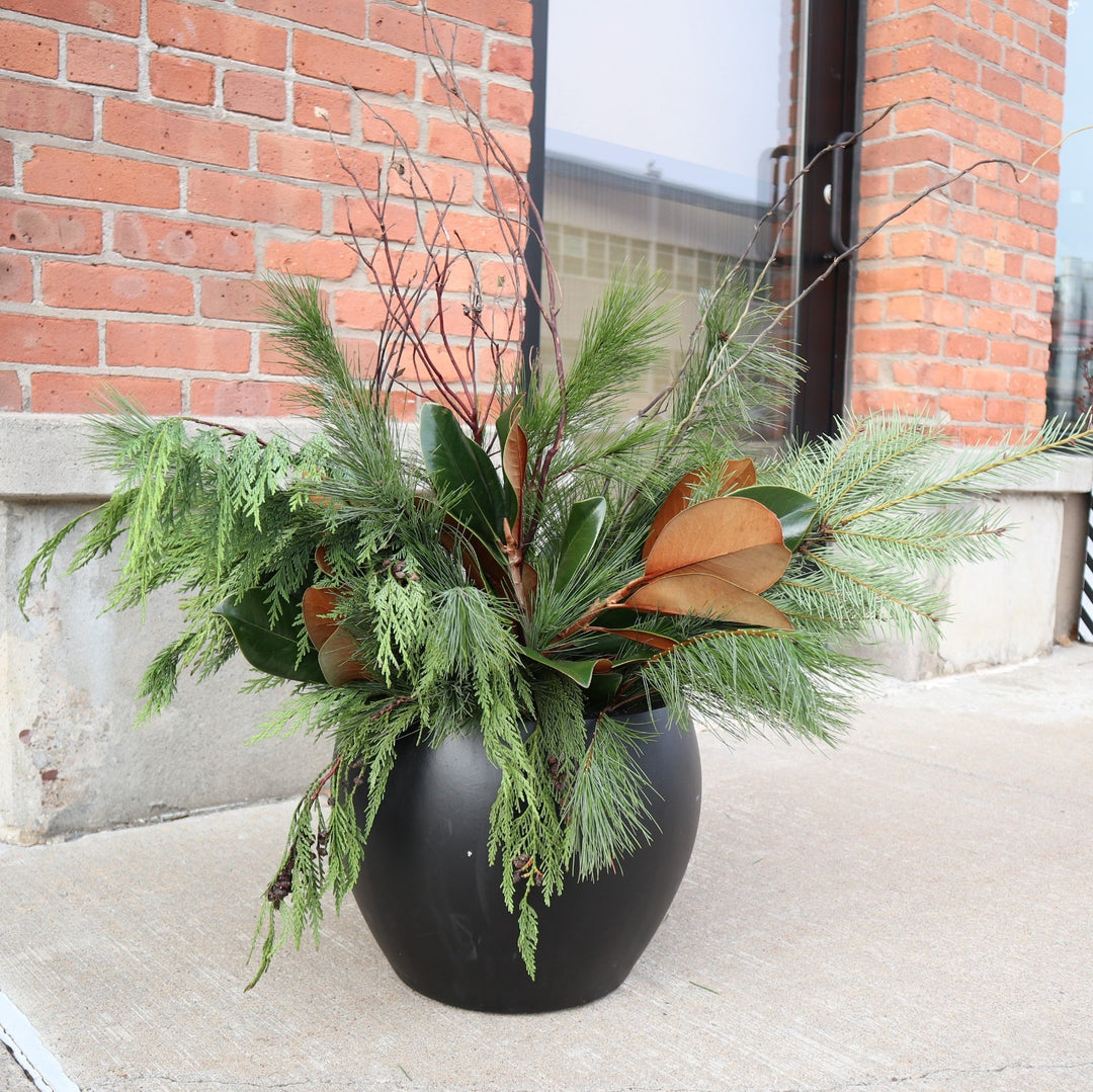 Potted arrangement with evergreens, magnolia, and branches. Photo taken outside on a sidewalk next to a brick wall.