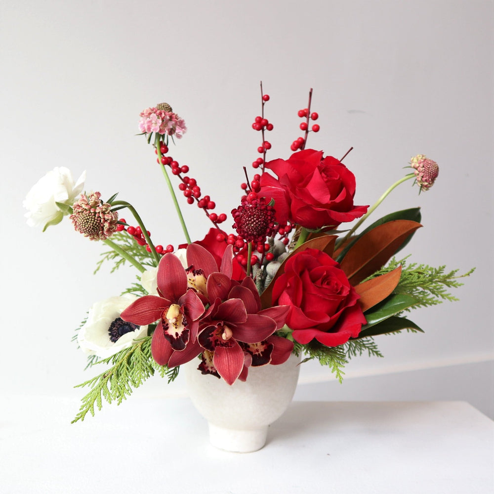 Arrangement in small white vessel, with red roses, cymbidium, scabiosa, ilex berries, evergreen, anemone, and magnolia.