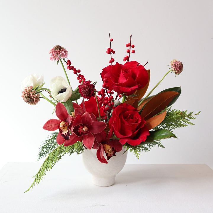 Arrangement in small white vessel, with red roses, cymbidium, scabiosa, ilex berries, evergreen, anemone, and magnolia.