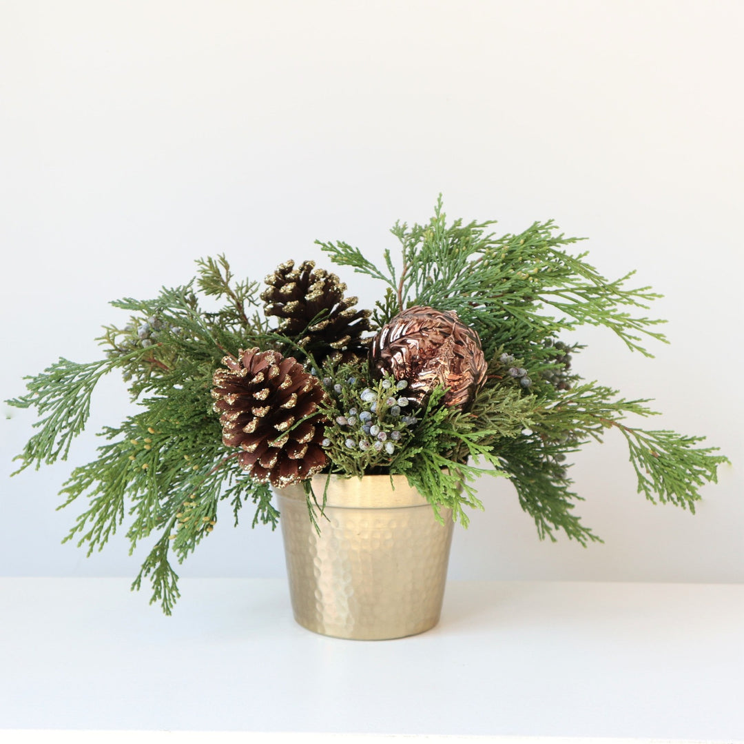 Arrangement in a gold metal vase, with evergreens, pinecones and an ornament.