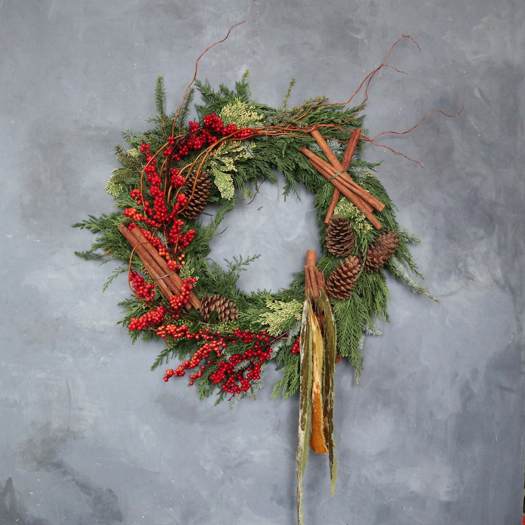 Organic evergreen wreath decorated with berries, branching, cinnamon sticks and a velvet style ribbon. Photo taken on gray backdrop.