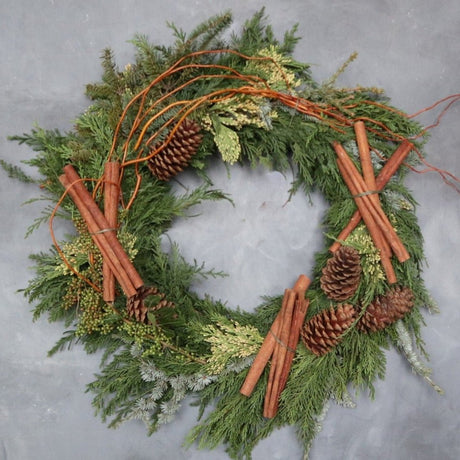 Evergreen wreath decorated with branching, different types of cedar, pinecones and cinnamon sticks. Photo taken on gray backdrop.