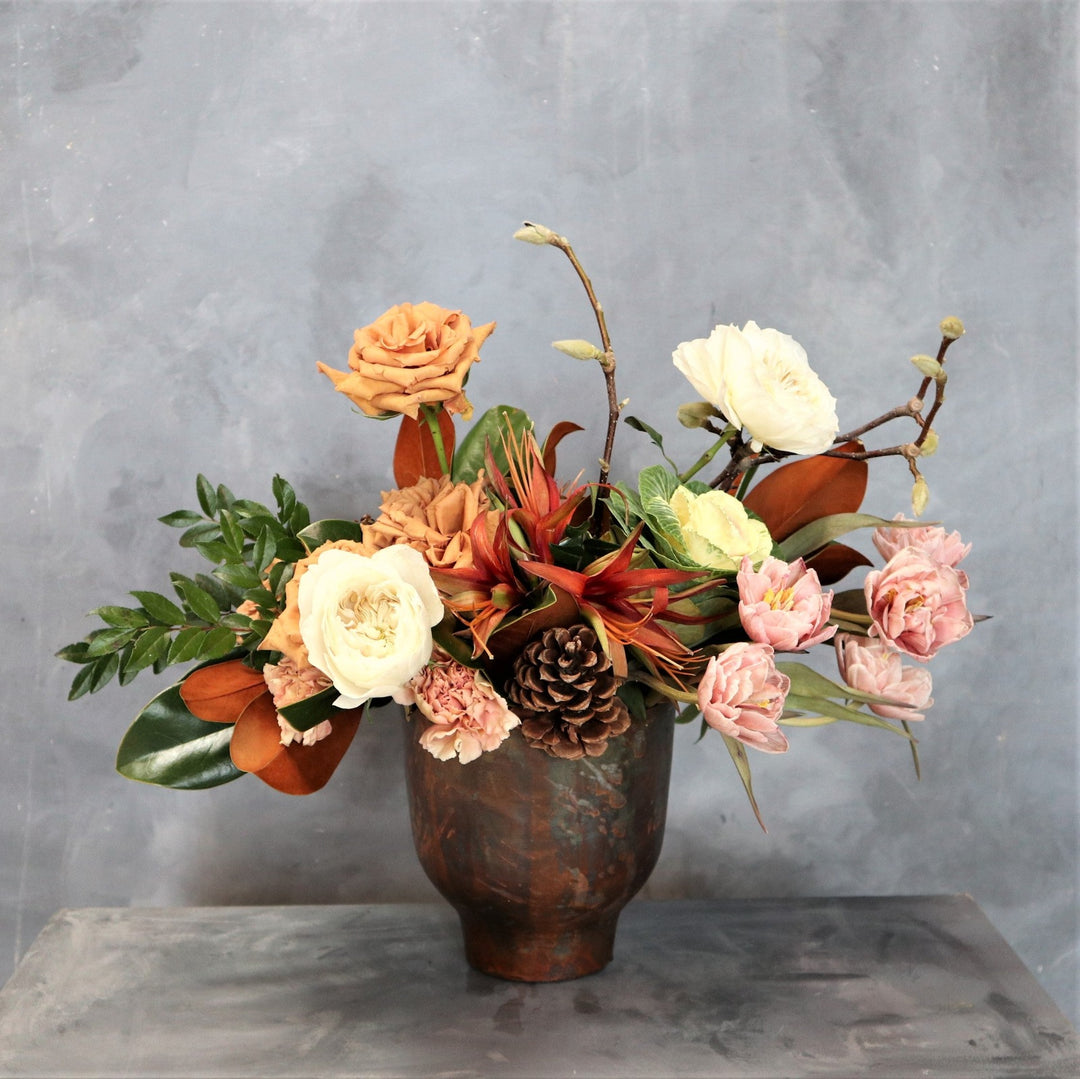 Arrangement in a brown metal vessel, filled with pink tulips, kale, carnations, toffee roses, amaryllis, magnolia branches, pinecones, greenery and magnolia. 