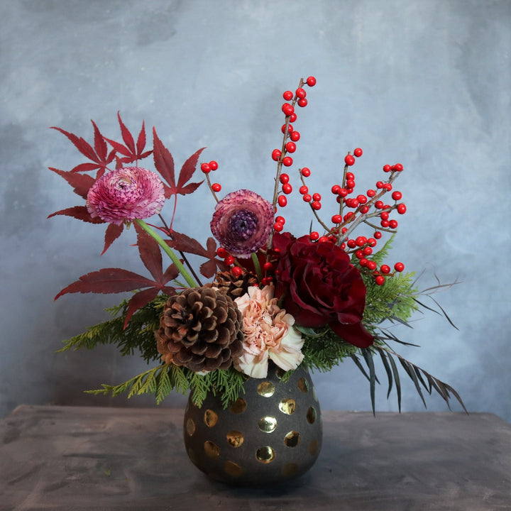 Vased arrangement in a polka dot vase, with carnation, red rose, pinecones, ranunculus, ilex berries and evergreen.