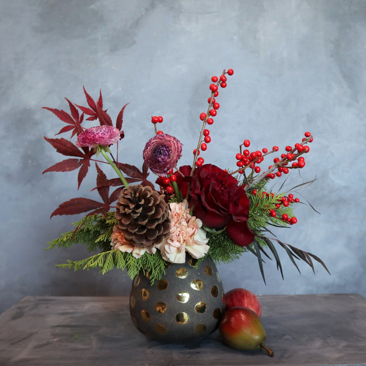 Vased arrangement in a polka dot vase, with carnation, red rose, pinecones, ranunculus, ilex berries and evergreen.