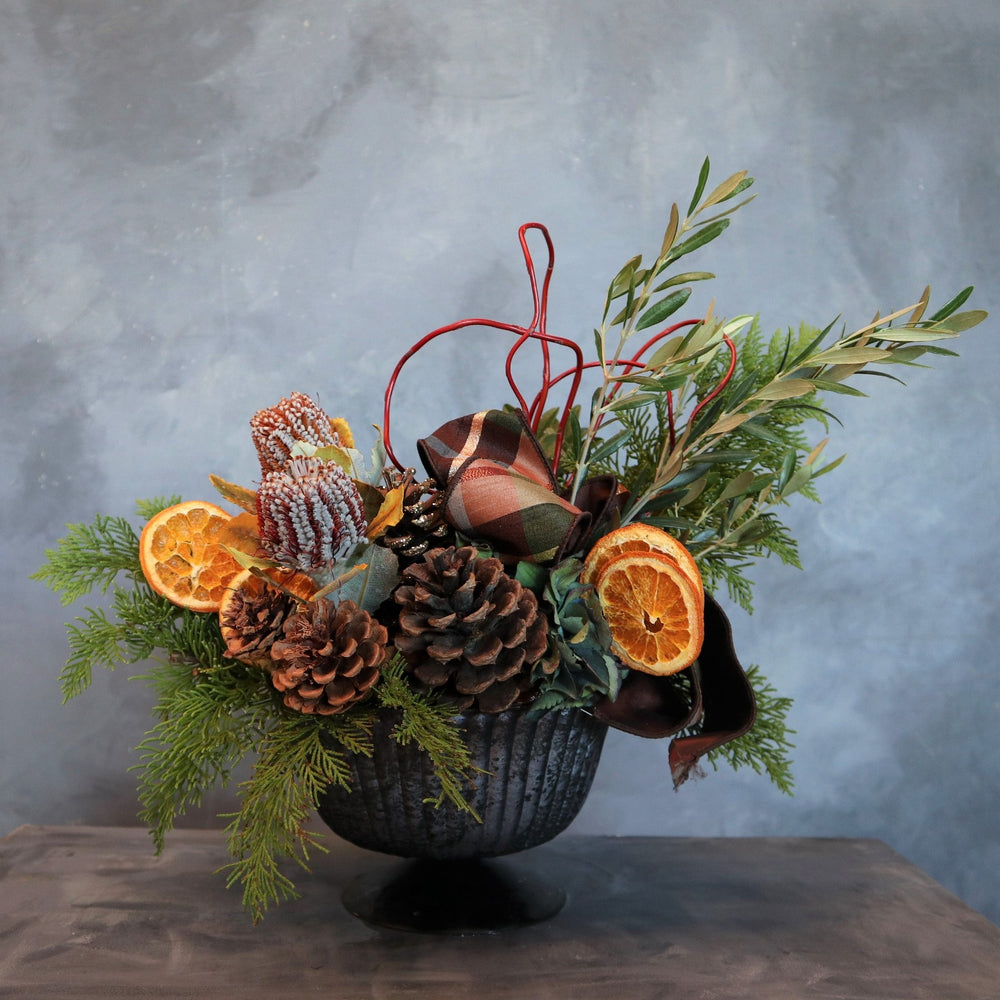 Arrangement in blue compote vessel, with pinecones, dried oranges, protea,evergreen, olive branches, curly willow and ribbon accents. Without fruit at the base.
