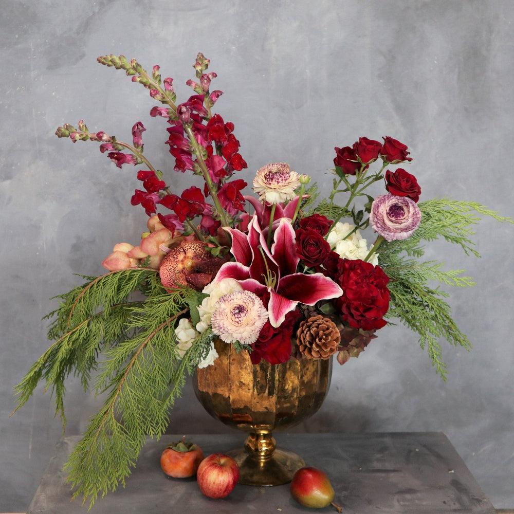 Arrangement in a gold mercury glass, with red lilies, red spray roses, white and purple ranunculus, orange vanda orchids, red roses, white hydrangea on a gray background.